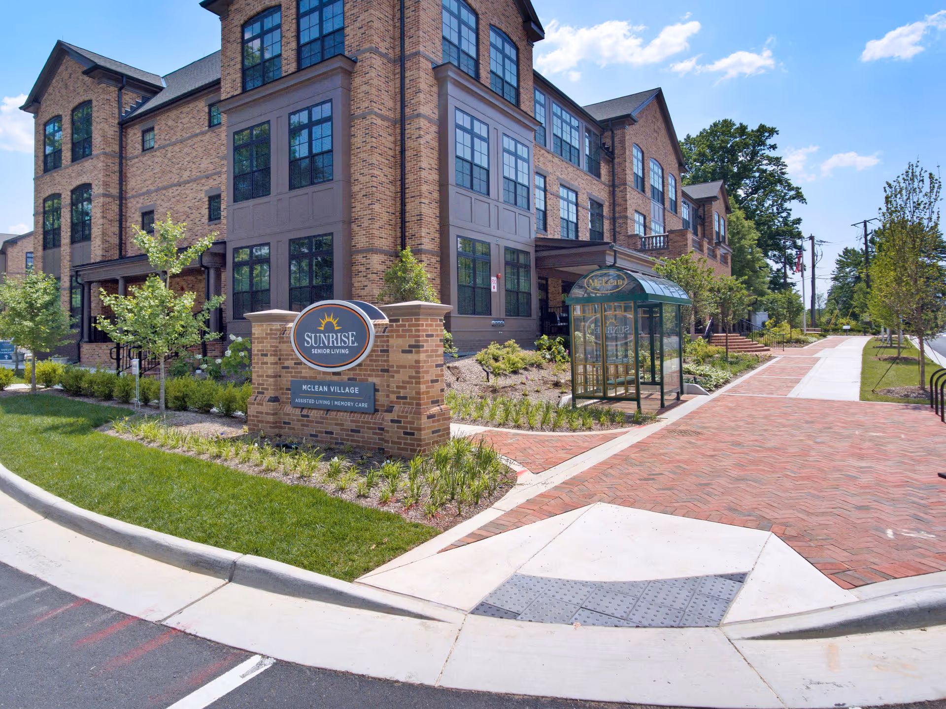 Exterior view of Sunrise of McLean Village senior living facility showing a large brick building with multiple windows, a landscaped area with small trees and bushes, a brick-paved sidewalk, and a bus stop shelter with a green roof. A sign in front reads 'Sunrise Senior Living McLean Village Assisted Living | Memory Care.'