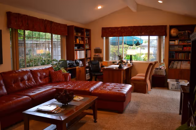 A cozy living room with a large red leather sectional sofa and matching ottoman. A wooden coffee table with magazines and a decorative bowl is in front of the sofa. The room has beige carpet and walls, with two large windows covered by red valances letting in natural light. There is a wooden desk with a black office chair and two additional chairs near the window. Bookshelves filled with books and decorative items flank the desk area. Outside the windows, greenery and a patio umbrella are visible.