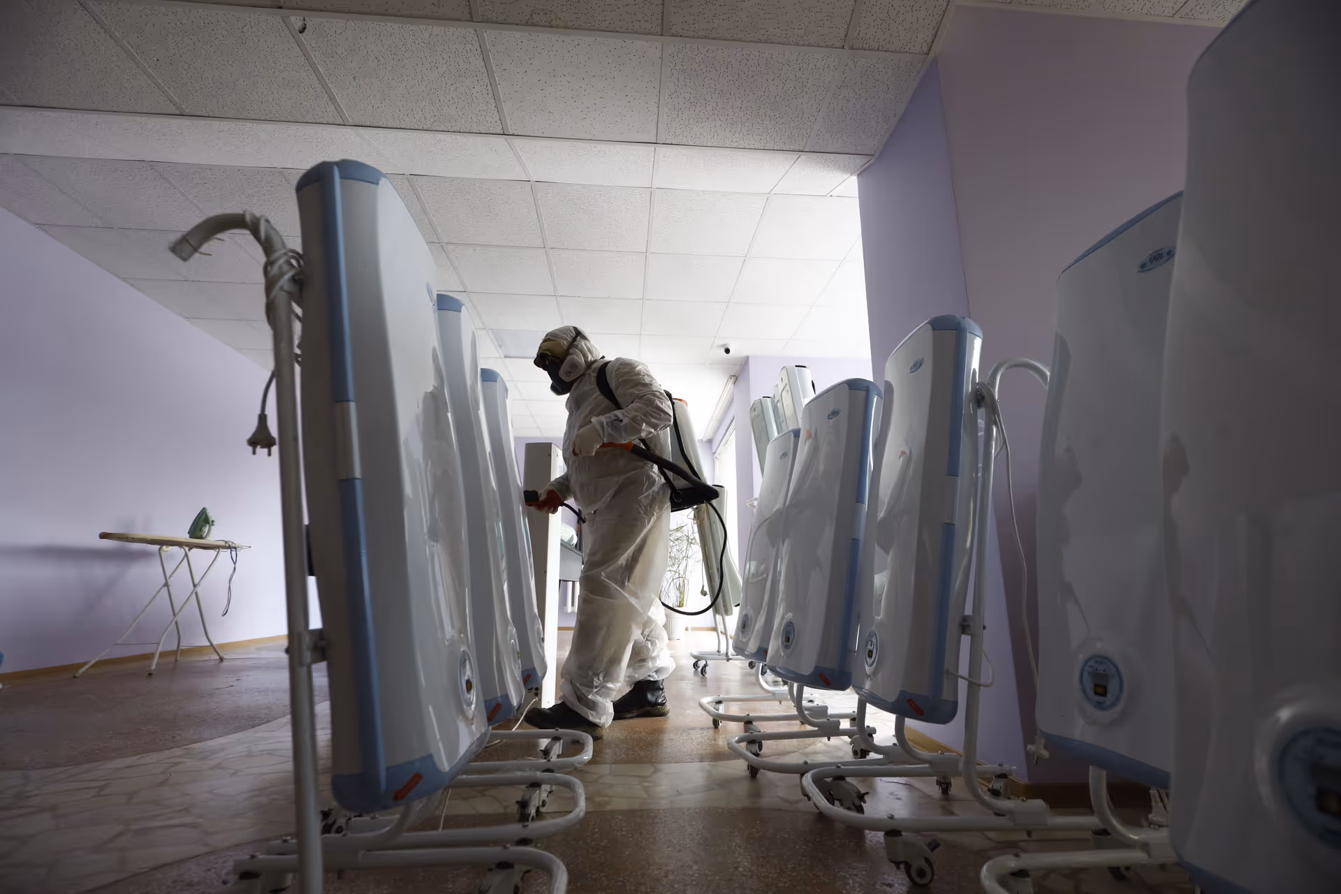 A worker in full PPE disinfects a row of medical devices inside a purple-walled facility.