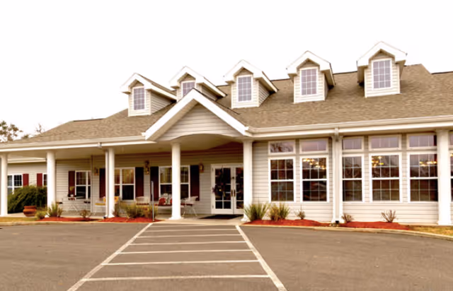 Front exterior view of a single-story building with a covered entrance supported by white columns, multiple windows, and dormer windows on the roof. There is a paved driveway with marked parking spaces leading up to the entrance.