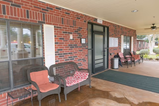 Covered brick entrance with cushioned wicker chairs, a glass door, and a welcome mat.