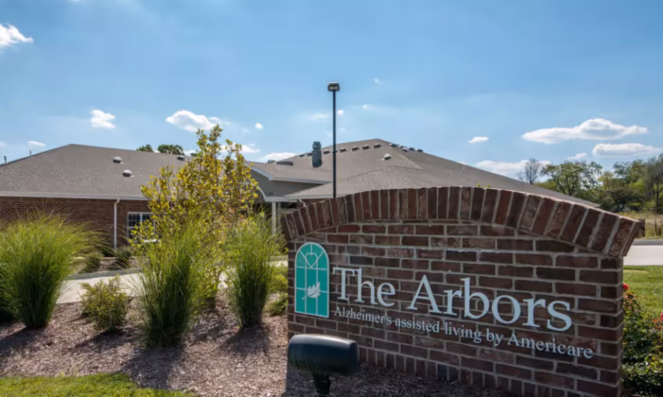 Exterior view of The Arbors at Westbrook Terrace, an Alzheimer's assisted living facility, showing a brick sign with the facility's name and logo in front of a landscaped area with bushes and a building in the background under a blue sky with some clouds.