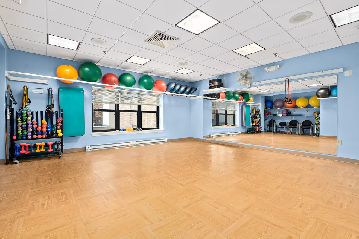 A spacious exercise room with light blue walls and wood-patterned flooring. The room features a large wall mirror, exercise balls in various colors on wall-mounted racks, and a rack of colorful dumbbells and resistance bands. There are windows with blinds letting in natural light and a ceiling with recessed lighting panels.
