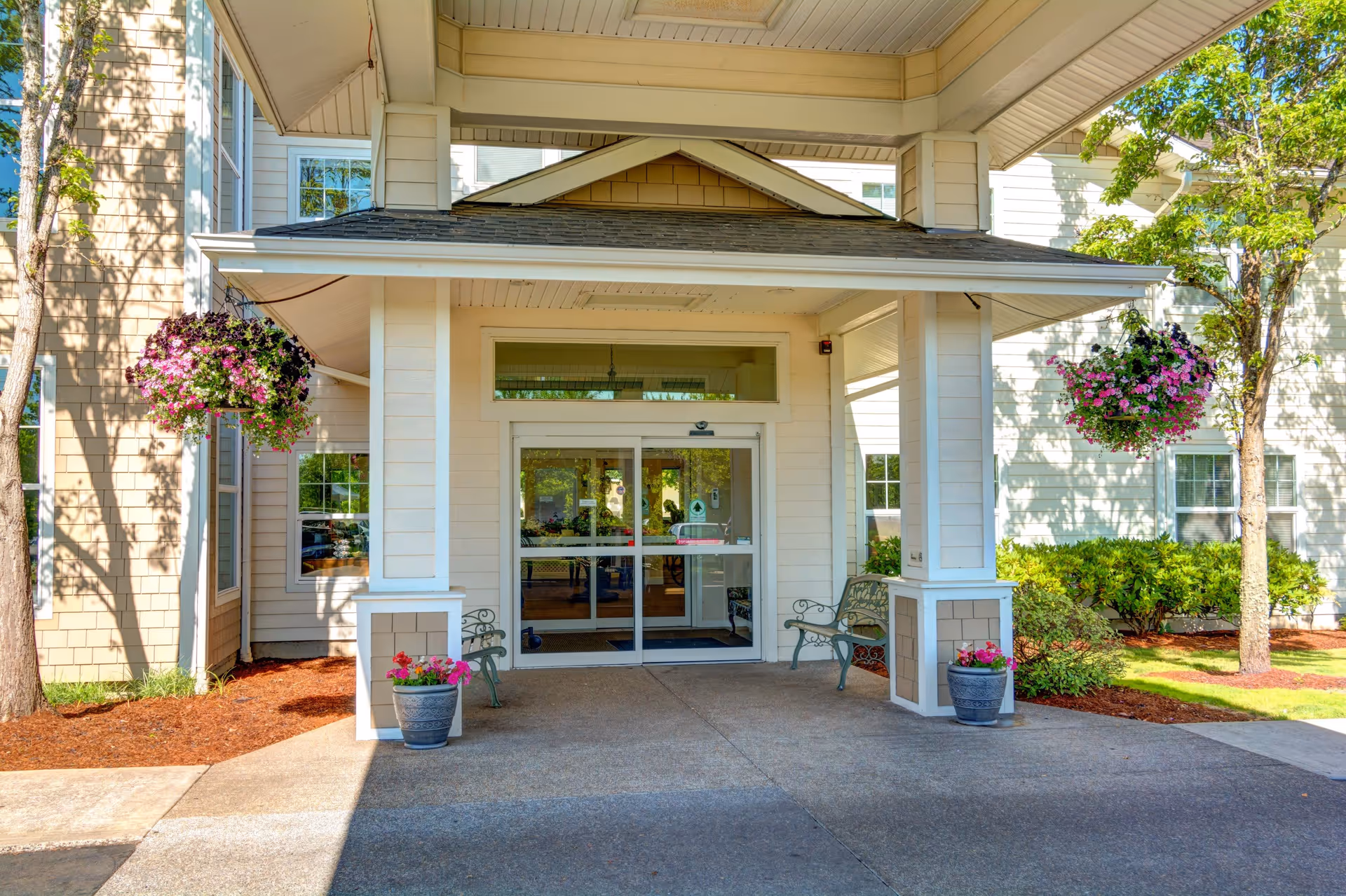 Entrance of Vineyard Heights Assisted Living facility with a covered porch, two benches on either side, hanging flower baskets, potted plants, and glass double doors leading inside.
