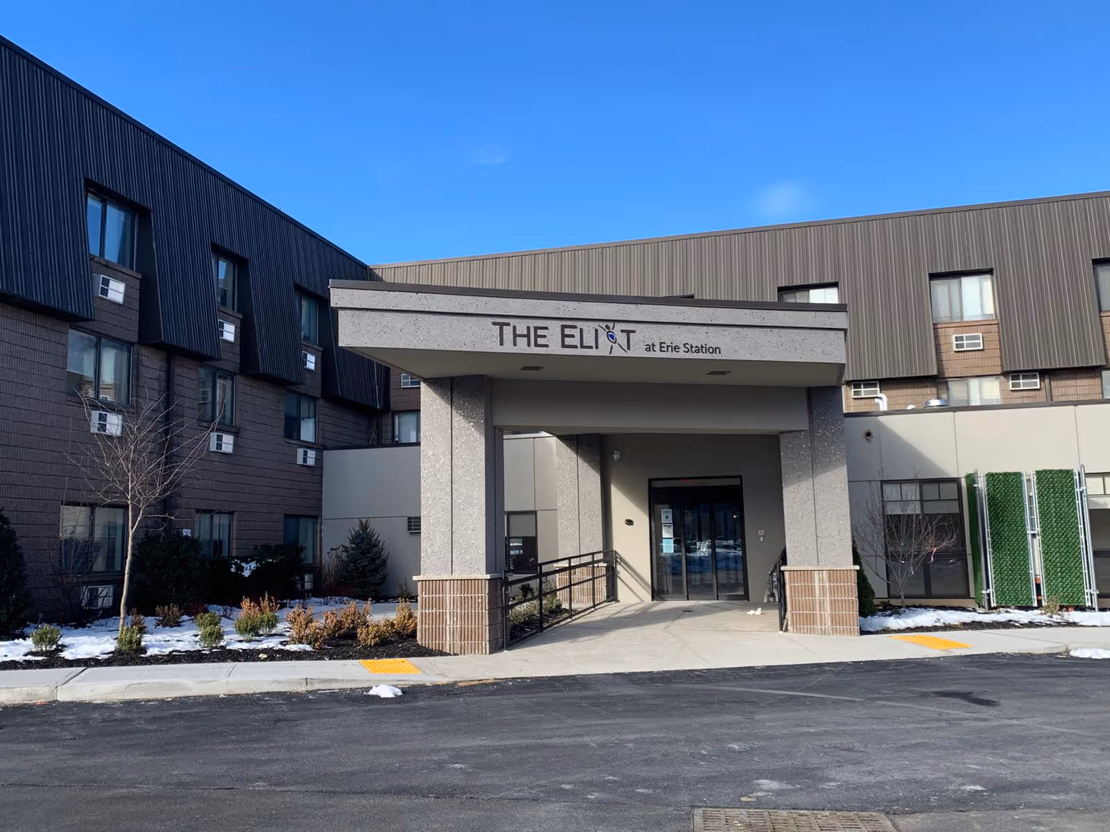 Exterior front entrance of The Eliot at Erie Station senior living facility with a covered drop-off area, surrounded by a multi-story building and some landscaping with patches of snow on the ground.