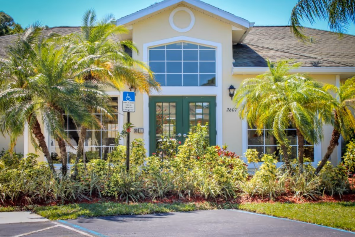 Front exterior view of a single-story building with a peaked roof, large windows, and double green doors. The entrance is surrounded by lush landscaping including palm trees and various shrubs. There is a handicap parking sign and a marked parking space in front of the building.