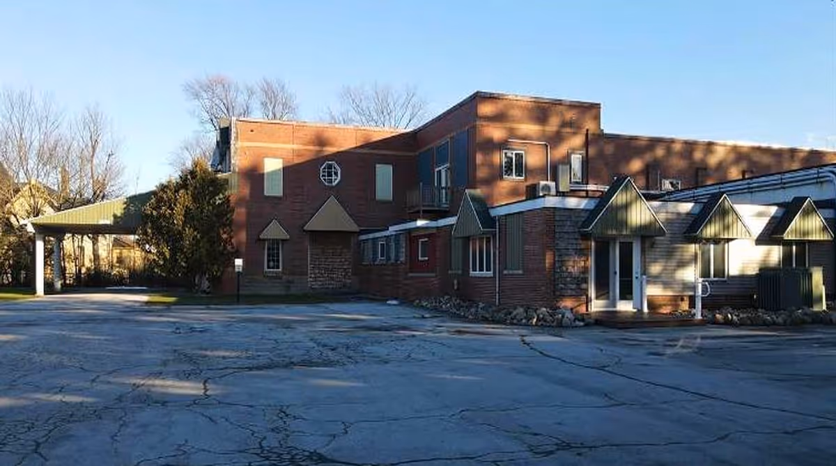 Exterior view of a brick and siding building with multiple windows and a covered entrance, surrounded by bare trees and an empty cracked parking lot under a clear sky.