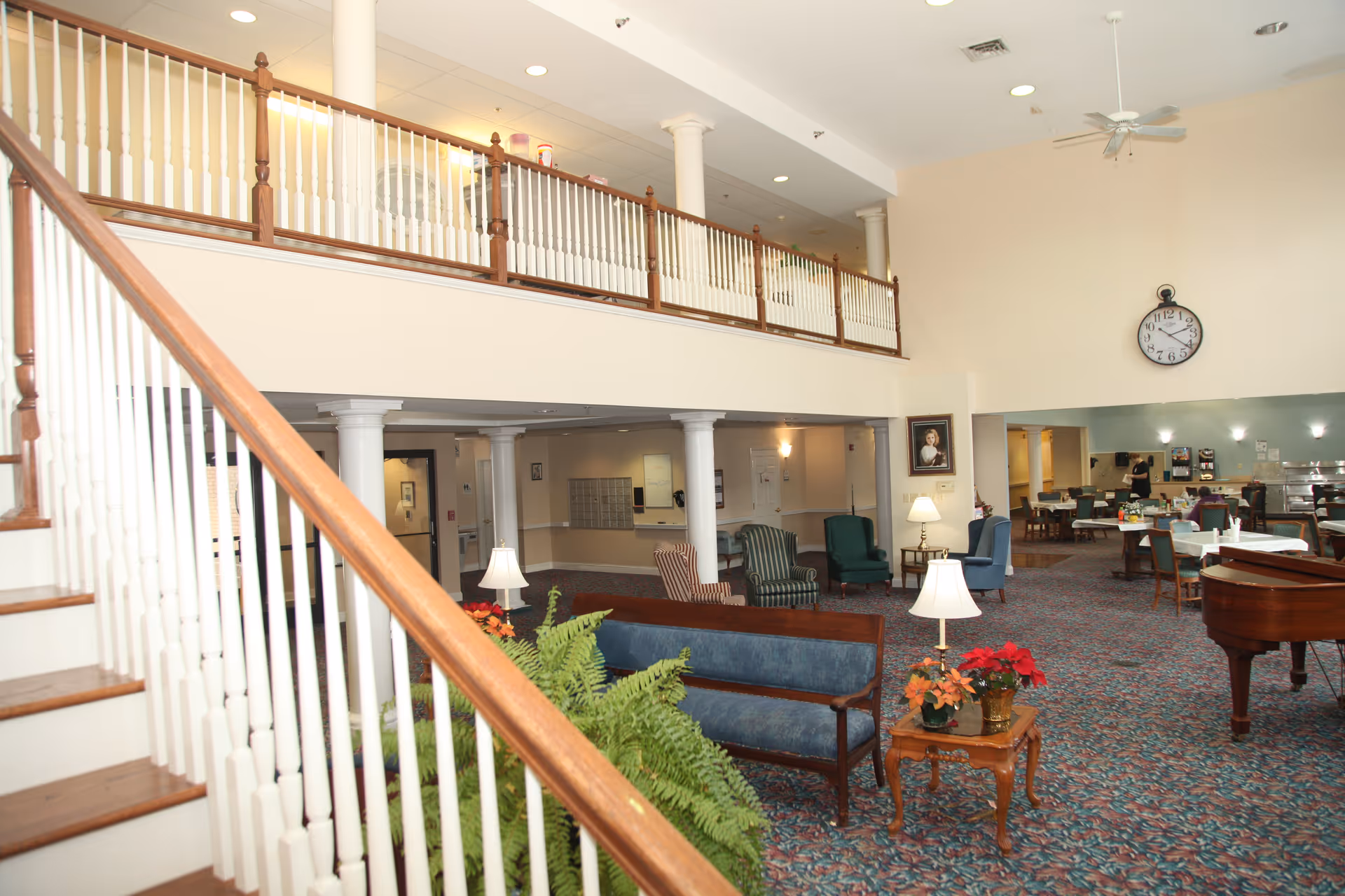 Spacious senior living facility common area with a staircase on the left, blue cushioned wooden benches, several armchairs, a small wooden table with potted plants, a grand piano on the right, and a dining area in the background. The room has high ceilings with recessed lighting and a large clock on the wall.