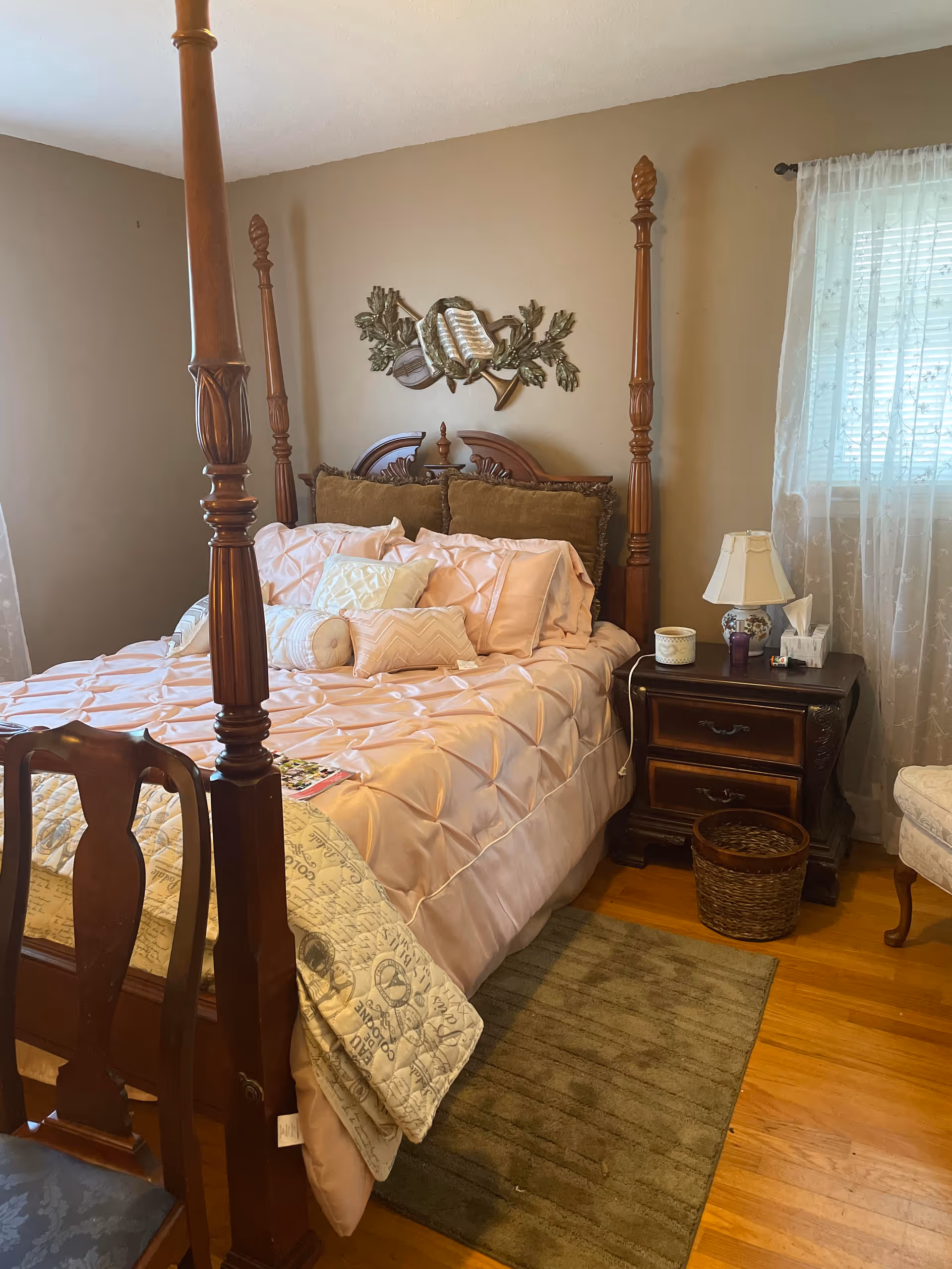 Four-poster bed with pink bedding, wooden nightstand, and sheer curtains over the window.