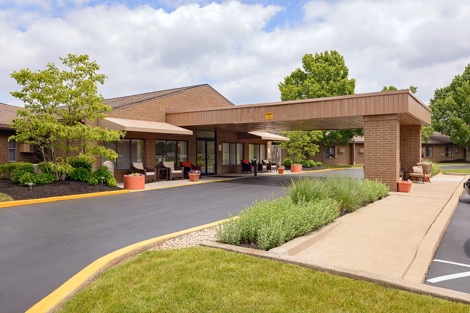 Covered porte-cochere entrance of a brick nursing care facility with seating, planters, and landscaped grounds.