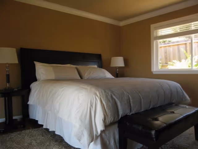 A bedroom with a large bed featuring white bedding and pillows, flanked by two dark wooden nightstands each with a lamp. There is a window on the right side letting in natural light, and a black cushioned bench at the foot of the bed. The walls are painted beige and the floor is carpeted.
