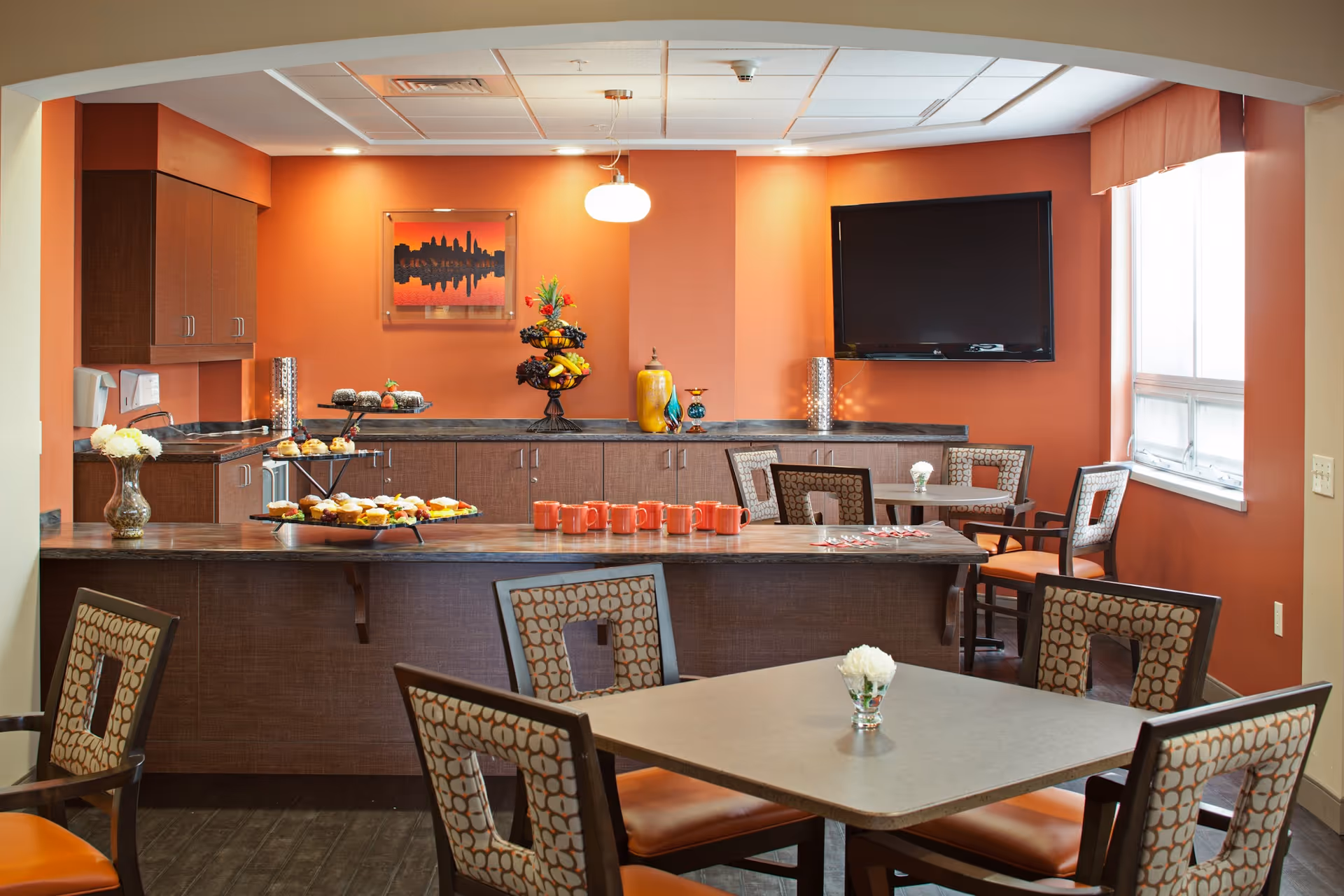A cozy dining area with orange walls featuring a countertop with snacks and orange mugs, several tables and chairs with patterned upholstery, a flat-screen TV mounted on the wall, a window letting in natural light, and decorative items including a vase with flowers and a fruit stand.