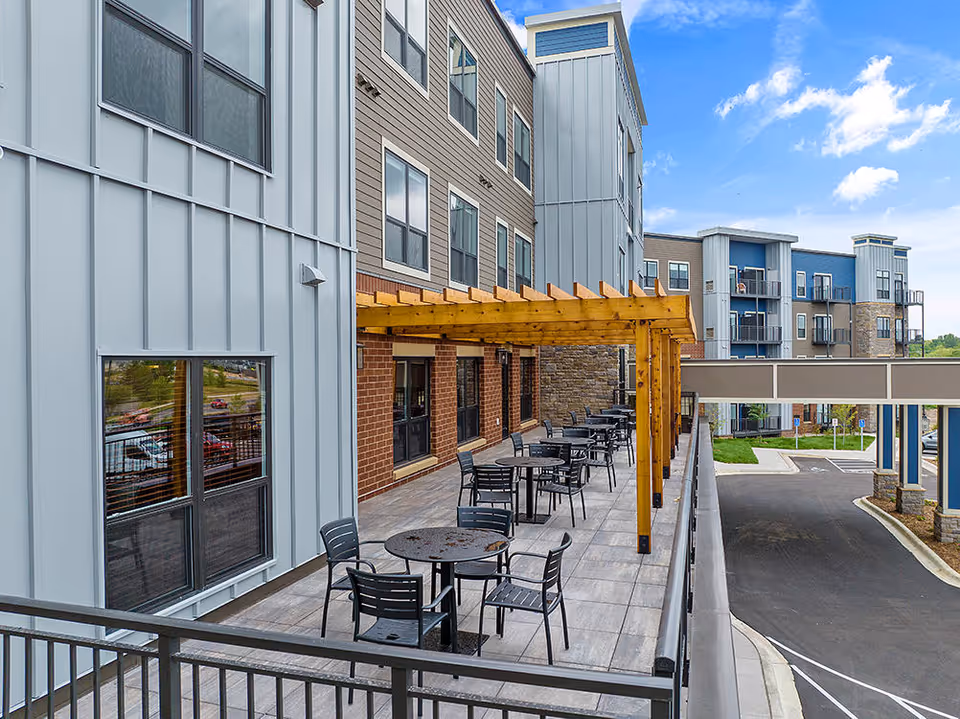 Outdoor patio area at The Meadows Senior Living with several black metal tables and chairs arranged under a wooden pergola attached to a multi-story building with mixed siding materials. The patio overlooks a parking area and other parts of the building under a partly cloudy blue sky.