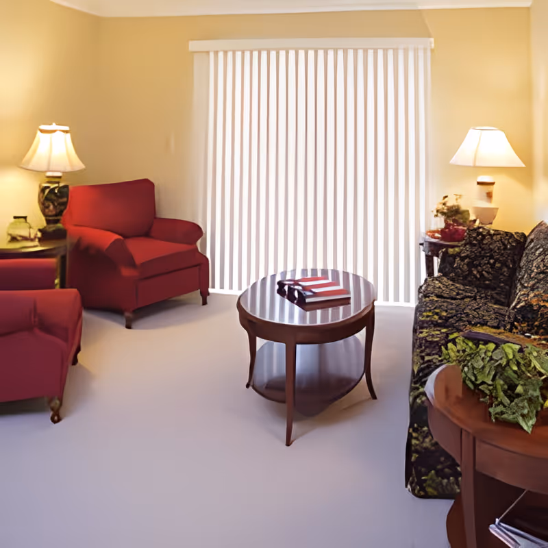 A cozy living room with two red armchairs, a patterned dark sofa, a wooden oval coffee table with books on it, two side tables with lamps, and vertical blinds covering a large window or sliding door.