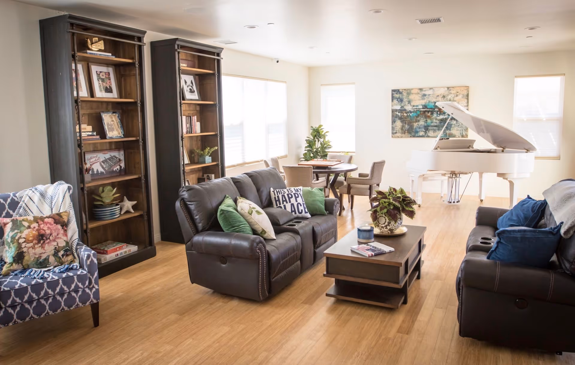 A bright living room with two dark leather sofas adorned with colorful pillows, a wooden coffee table with a plant and books, a blue patterned armchair with a floral pillow, two tall wooden bookshelves filled with books and decor, a round dining table with chairs near the windows, and a white grand piano against the far wall under a large abstract painting.