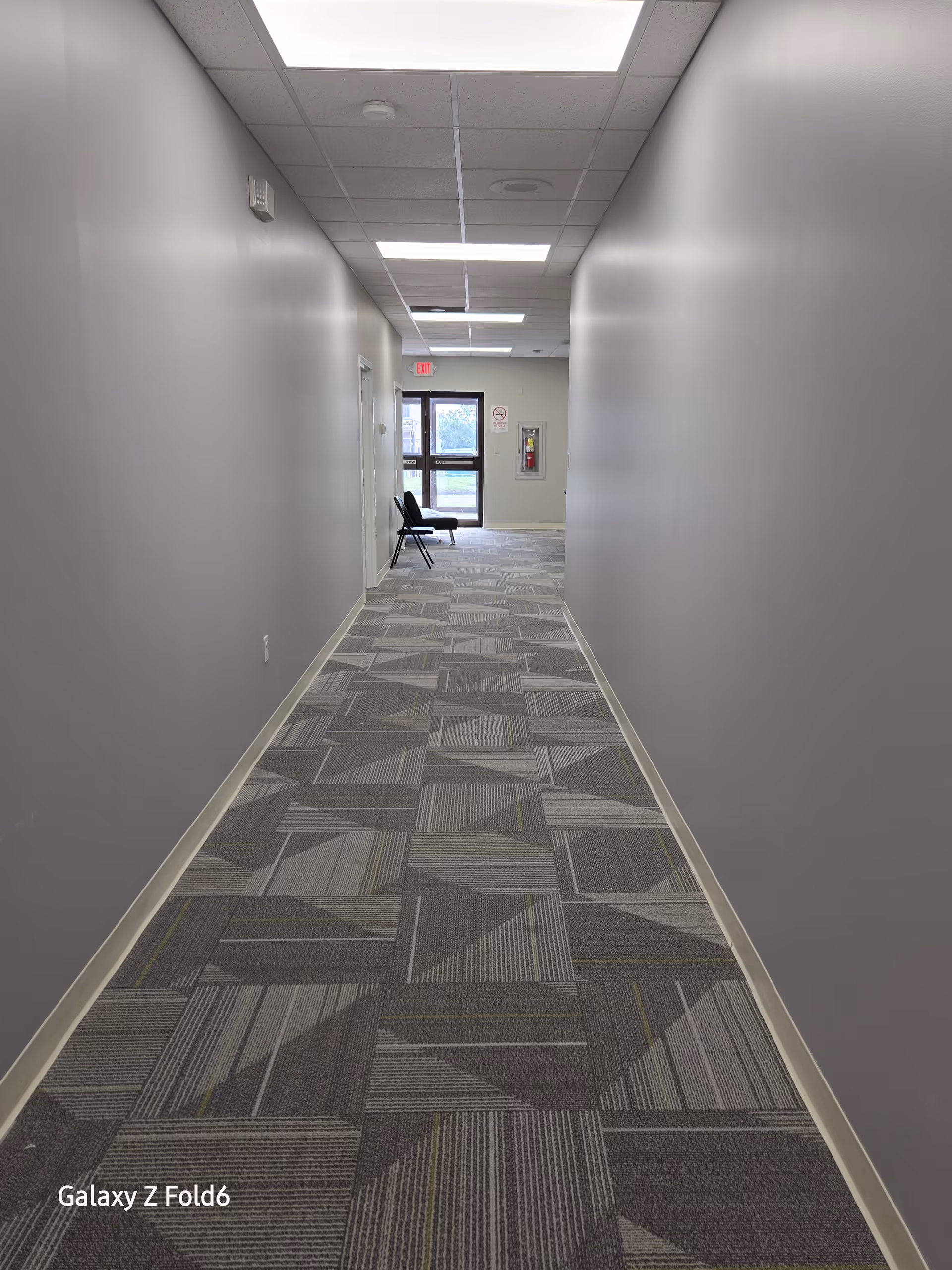 Long gray hallway in a senior living facility with carpet, lights, and a chair near exit.