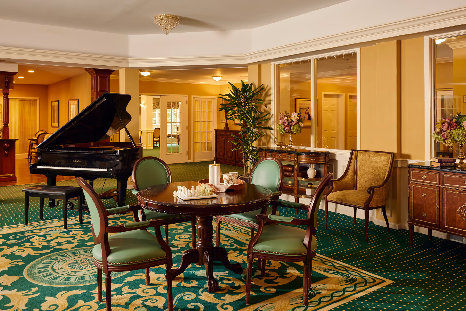 A cozy and elegant common area with a round wooden table surrounded by four green upholstered chairs. On the table, there is a chess set and a tray with a teapot and cups. In the background, there is a black grand piano with a matching bench, a large green plant, and wooden cabinets with floral arrangements. The room features green patterned carpet, yellow walls, and large windows with white trim.