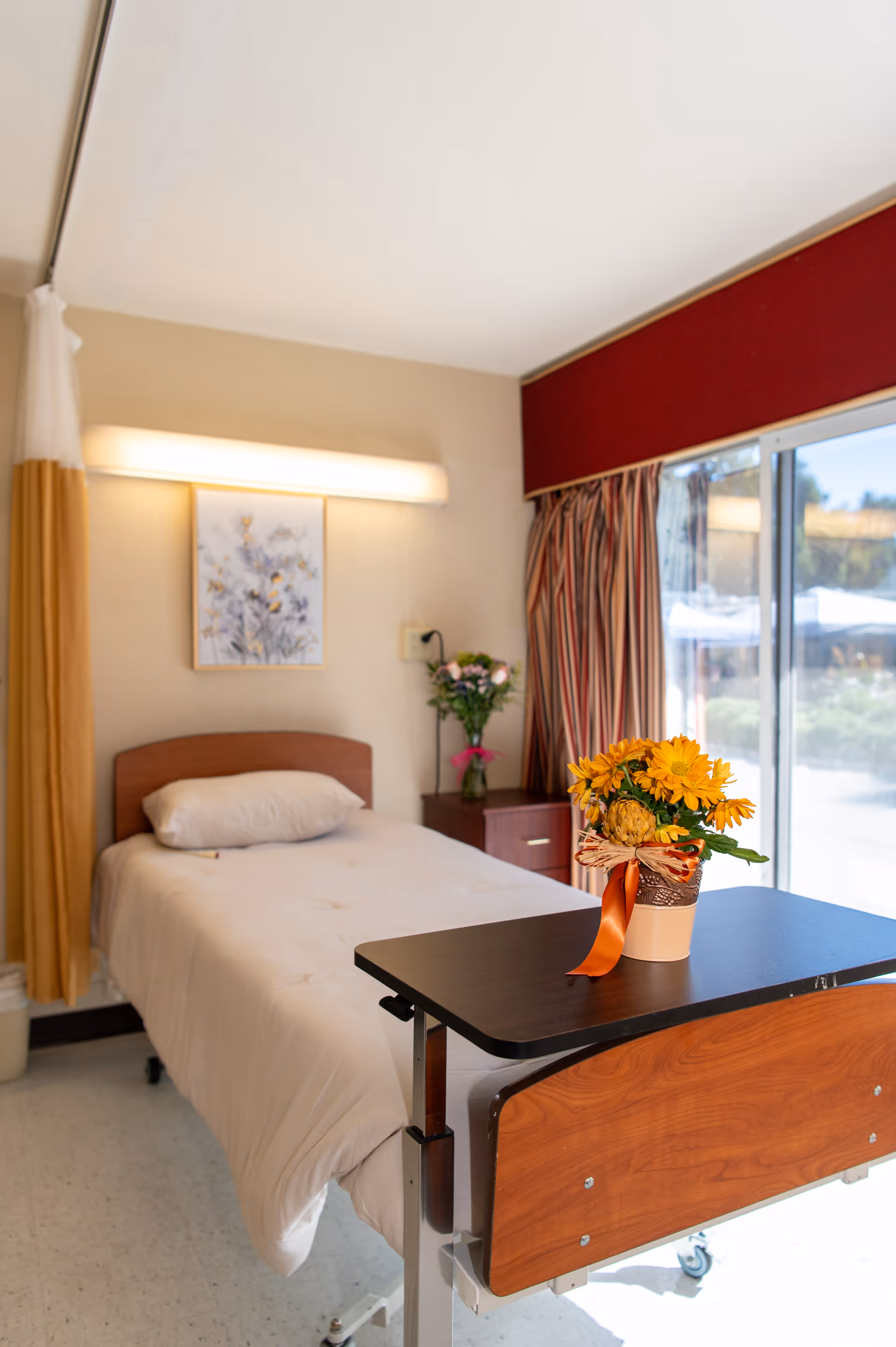 A hospital-style bedroom in a senior living facility with a bed, flowers, and a window view.