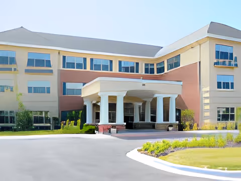 Exterior view of a three-story senior living facility building with a covered entrance supported by white columns, surrounded by landscaped greenery and a paved driveway.