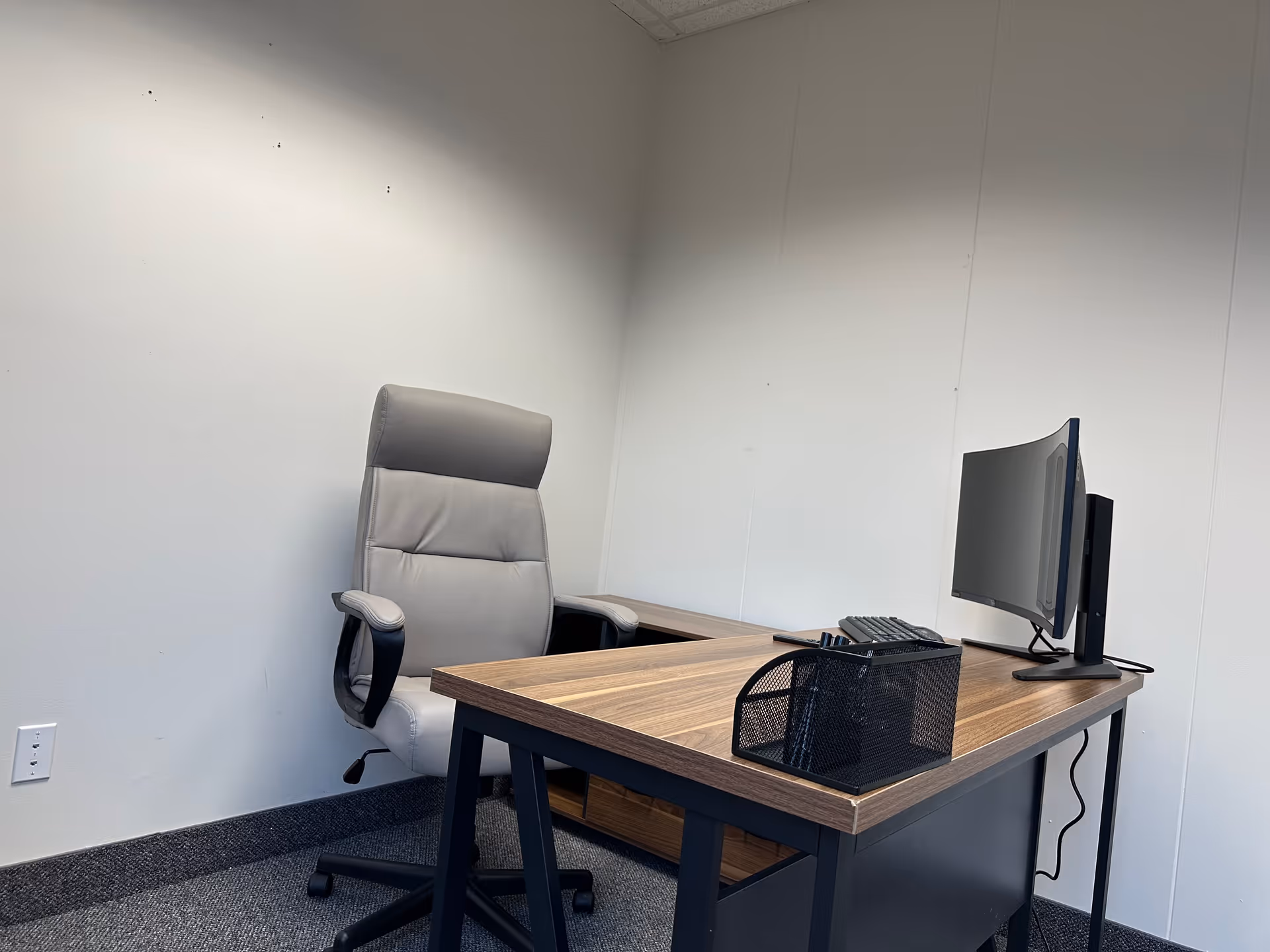 Office workspace with a wooden desk, computer, and gray ergonomic chair in a bare room.