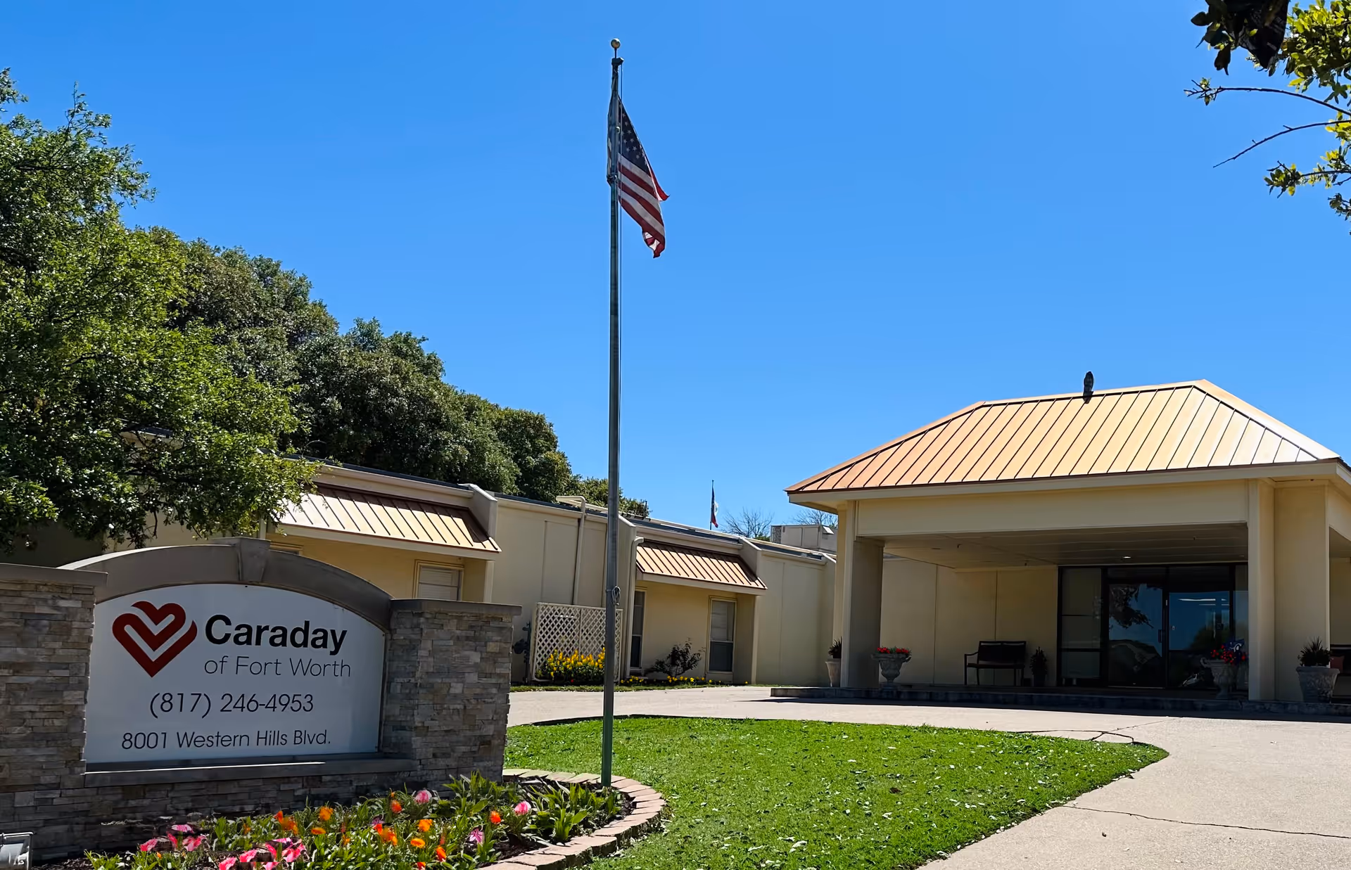 Front entrance of a Caraday assisted living building with a flagpole, landscaping, and a stone sign reading 'Caraday of Fort Worth'.
