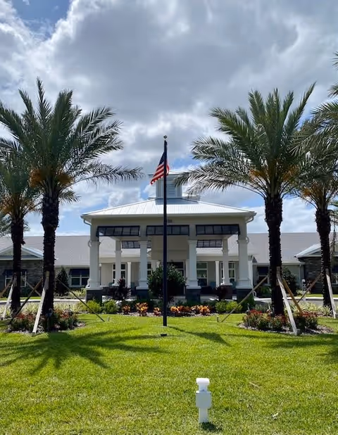 Front entrance of The Addison of Narcoossee with palm trees, an American flag on a pole, and a manicured lawn under a cloudy sky.