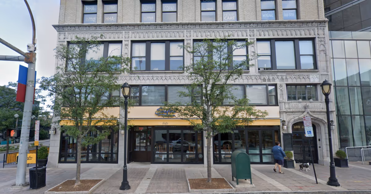 Urban building facade with large storefront windows, doorway, trees, and a person walking a dog.