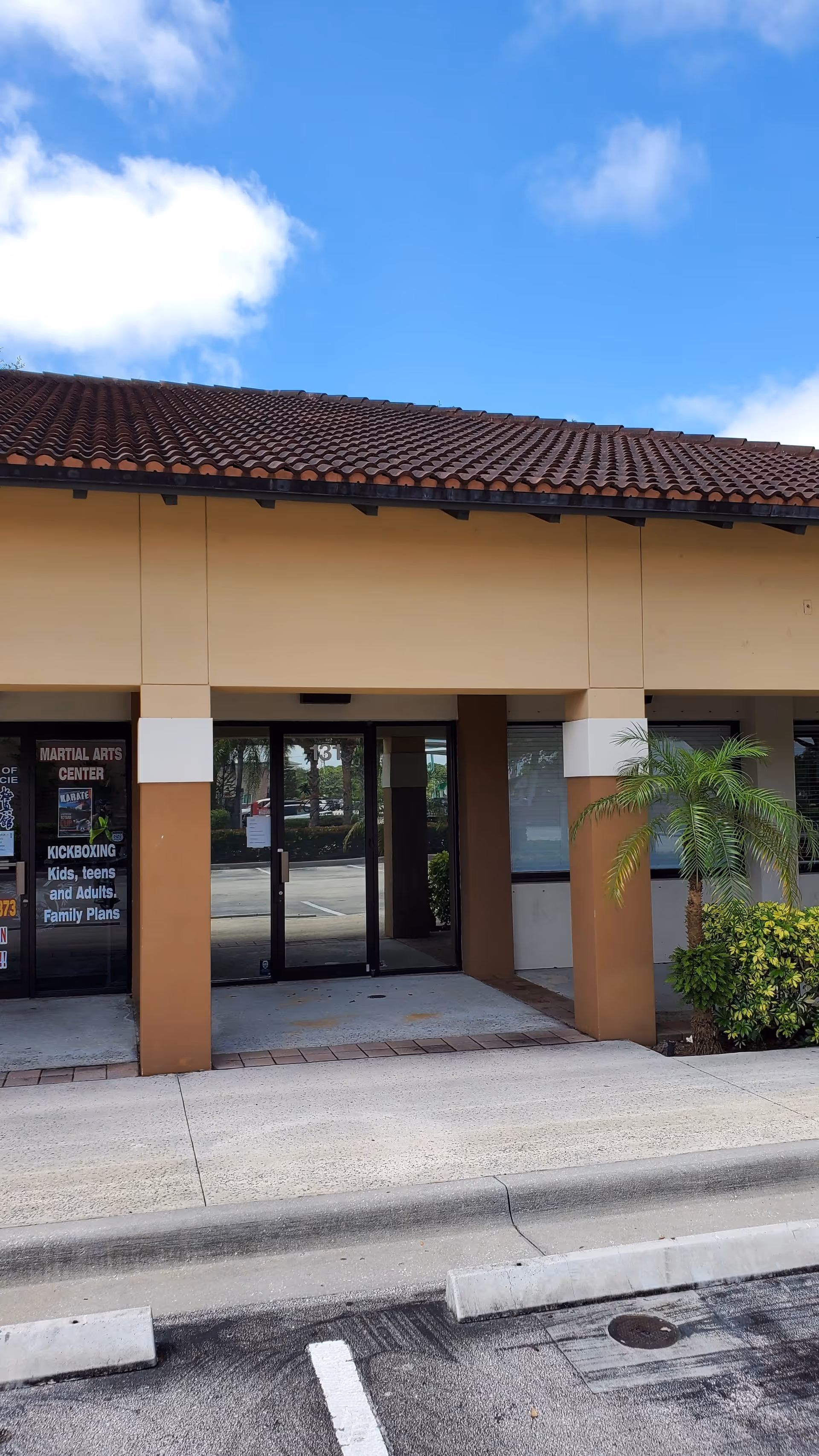 Exterior entrance of a tan building with tiled roof, glass doors, and a palm plant.