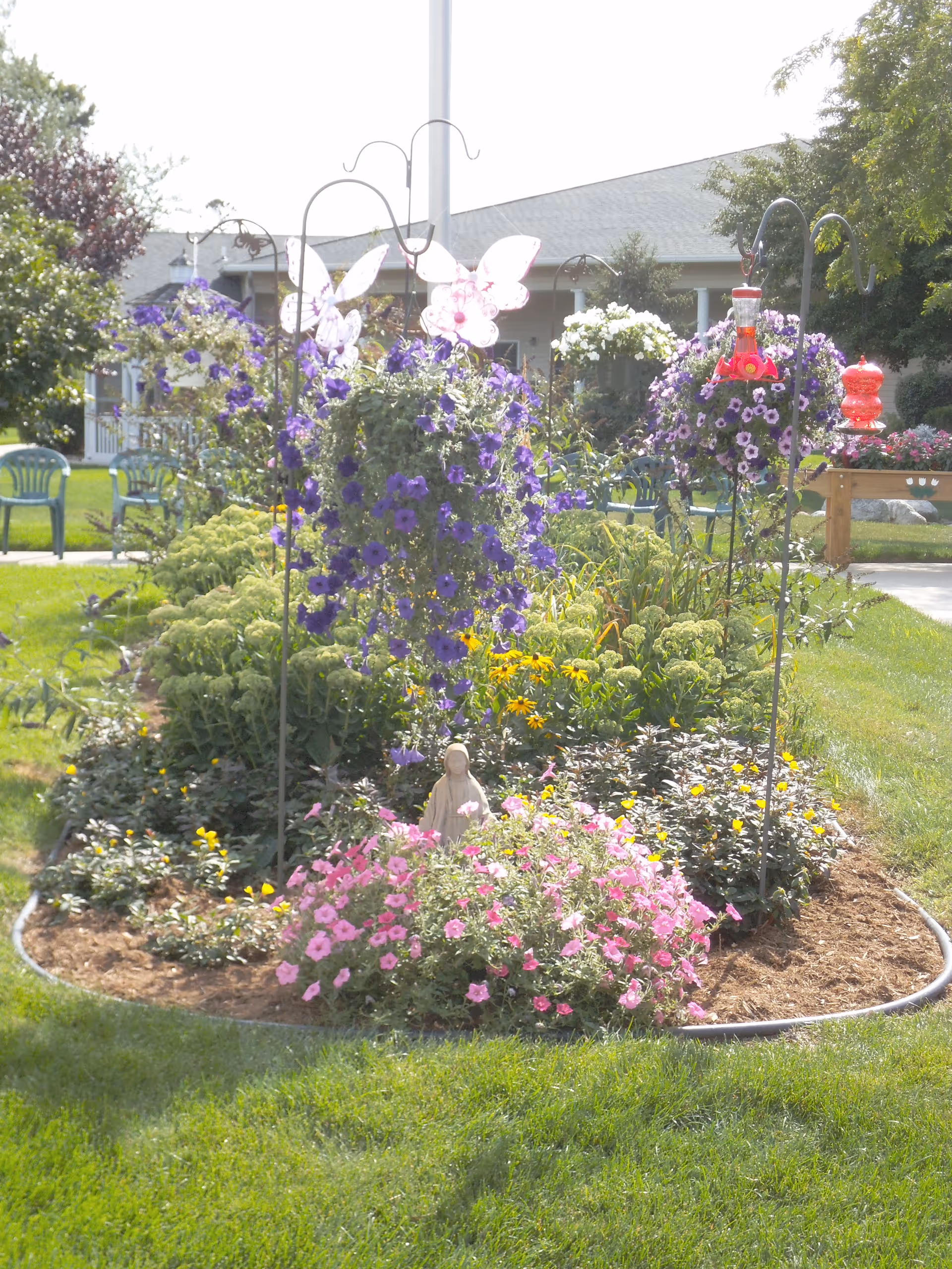 A landscaped outdoor garden bed with purple and pink flowers, hanging baskets, decorative butterfly ornaments and bird feeders in front of a building.