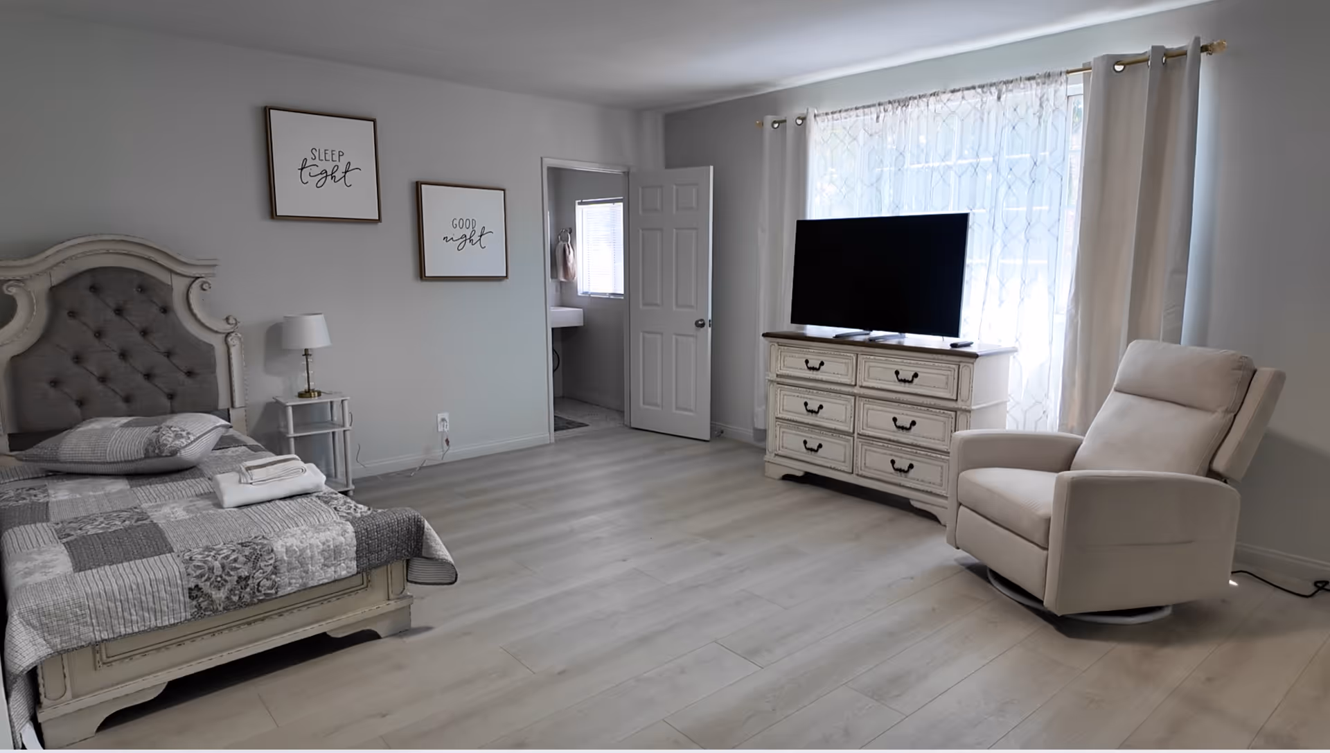 Gray-toned bedroom featuring a bed, dresser, TV, recliner, and large curtained window.