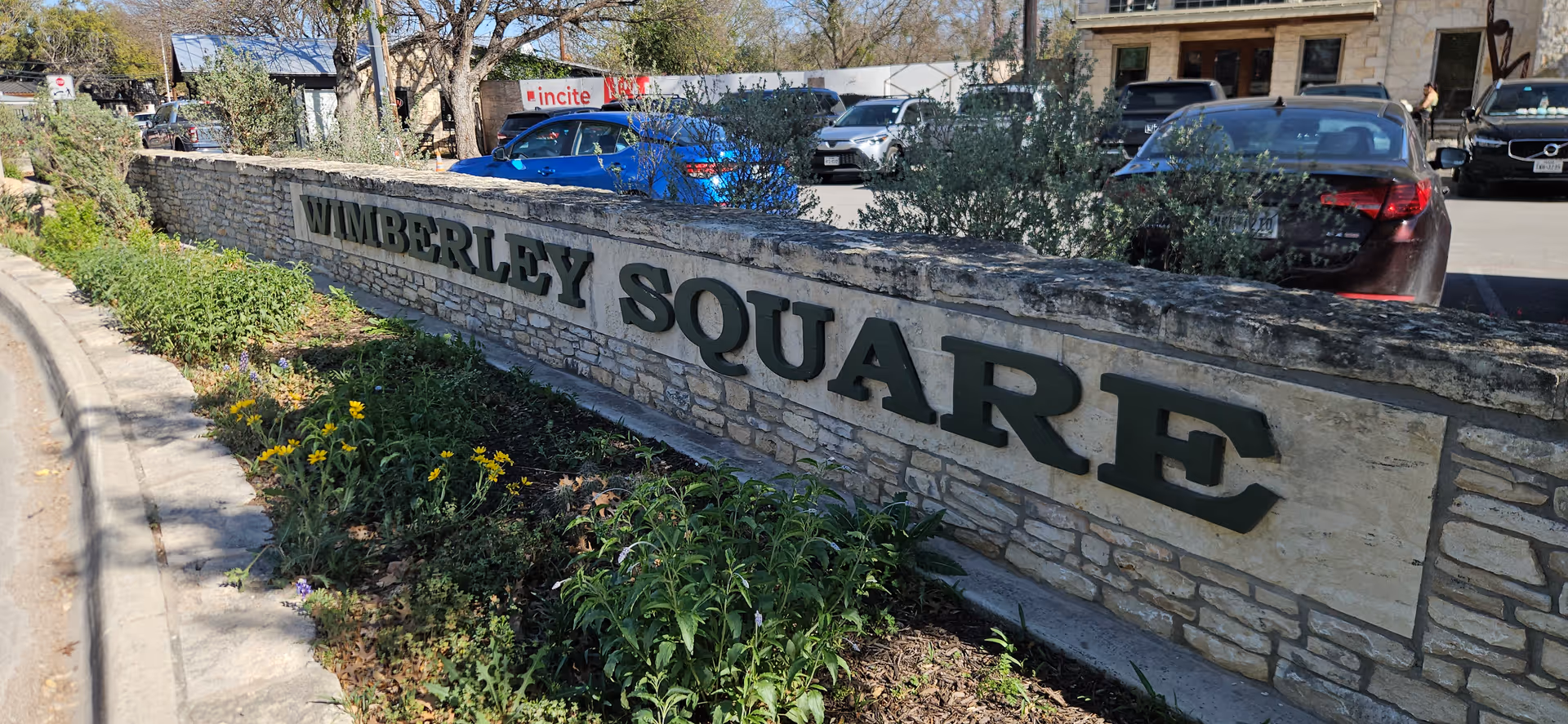 Exterior view of a stone sign reading Wimberley Square with cars behind.
