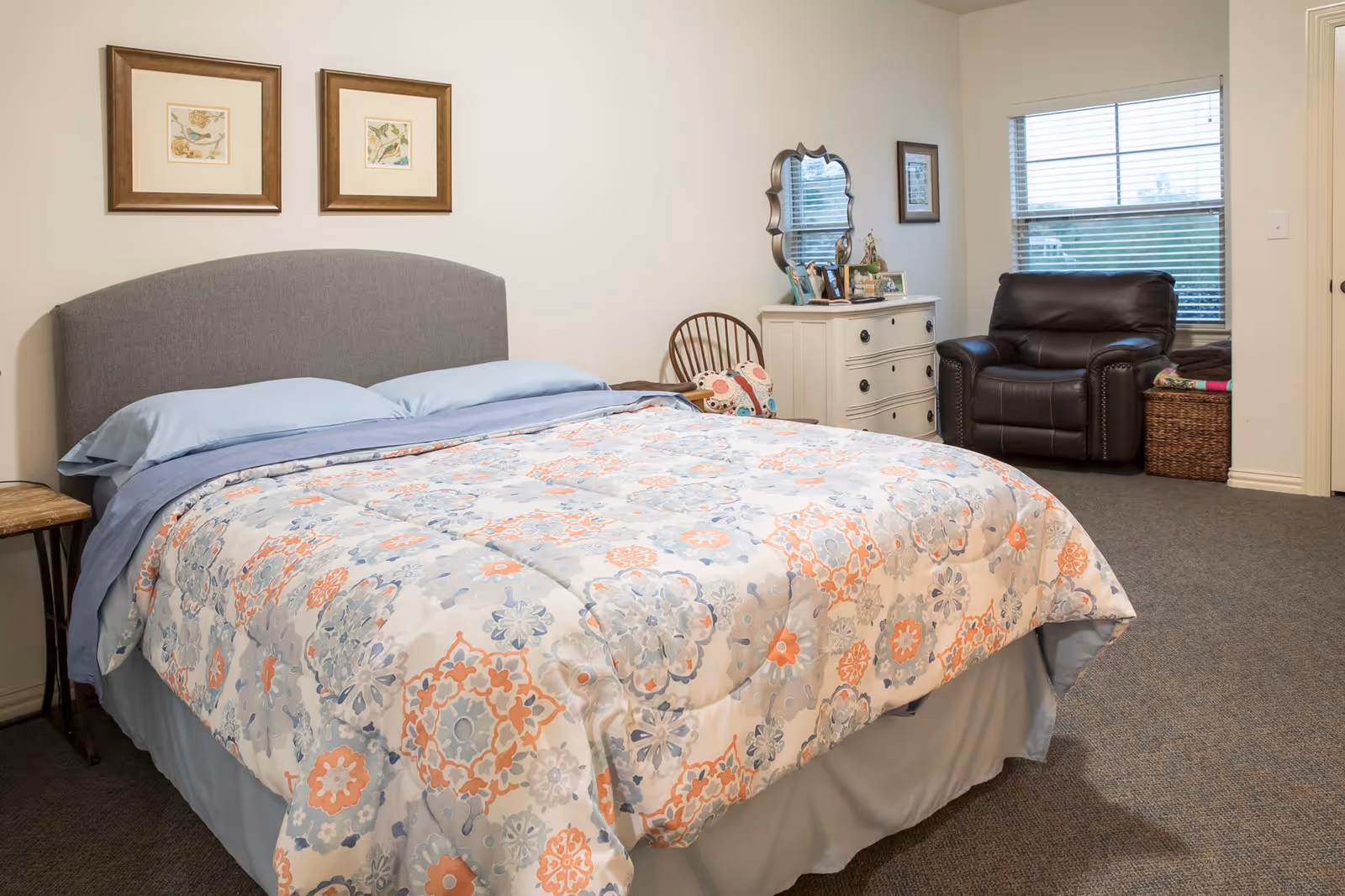 A bedroom with a bed covered in a floral patterned comforter in shades of blue, orange, and white. The bed has a gray upholstered headboard and blue pillows. On the left side of the bed is a small wooden side table. Against the far wall is a white dresser with a decorative mirror and framed photos on top. Next to the dresser is a wooden chair with a colorful cushion. In the corner near a window with blinds is a dark brown leather armchair and a woven basket. The room has beige walls and carpeted flooring.
