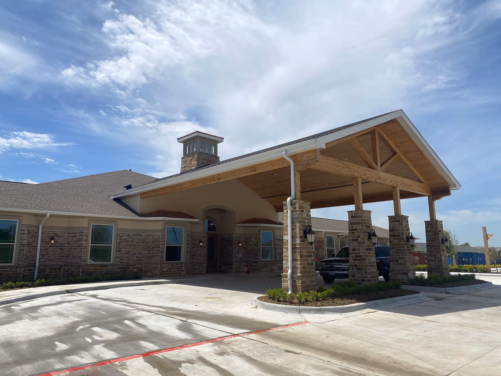 Exterior view of Village Green Memory Care Community Rockwall building with a covered entrance supported by stone pillars, a vehicle parked under the entrance, and a partly cloudy sky overhead.