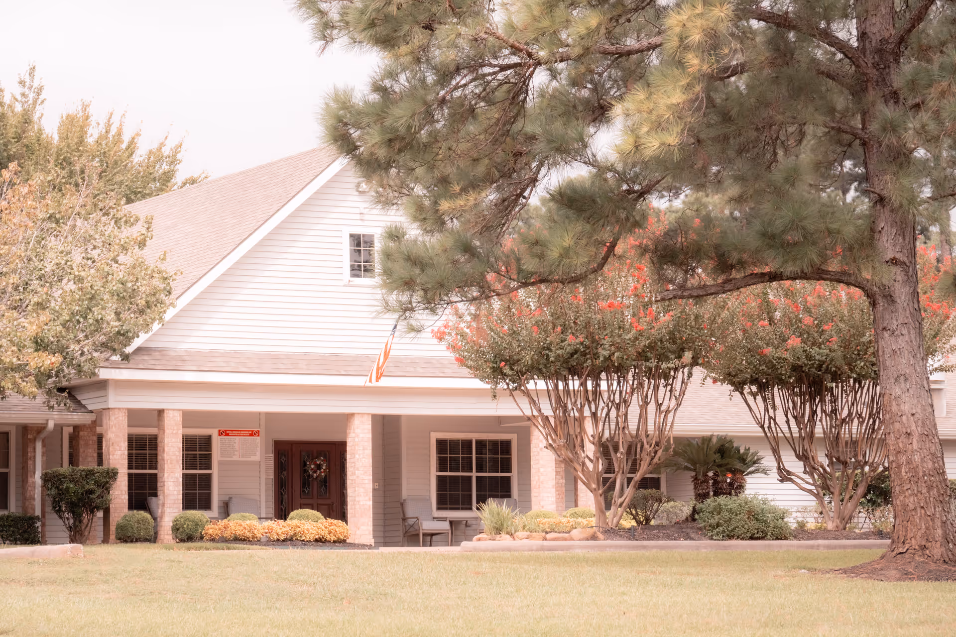 Front exterior view of a single-story building with a covered porch supported by brick columns, surrounded by well-maintained landscaping including bushes, trees, and a lawn. An American flag is displayed near the entrance.