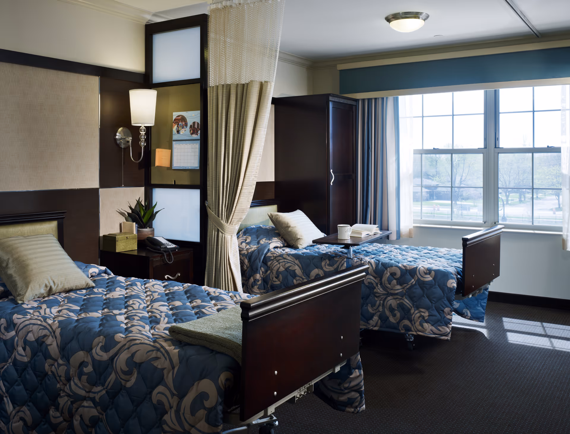 Two twin beds with blue patterned quilts occupy a senior living facility bedroom by a window.