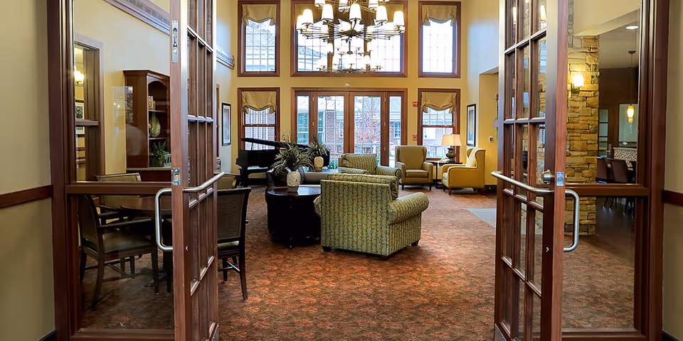 View through open wooden double doors into a spacious living room area with patterned carpet, multiple armchairs and sofas arranged around coffee tables, a grand piano near large windows with beige curtains, and a chandelier hanging from the high ceiling.