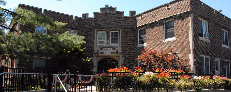 Two-story brick building with a decorative entrance, railing and colorful flowerbeds in front.