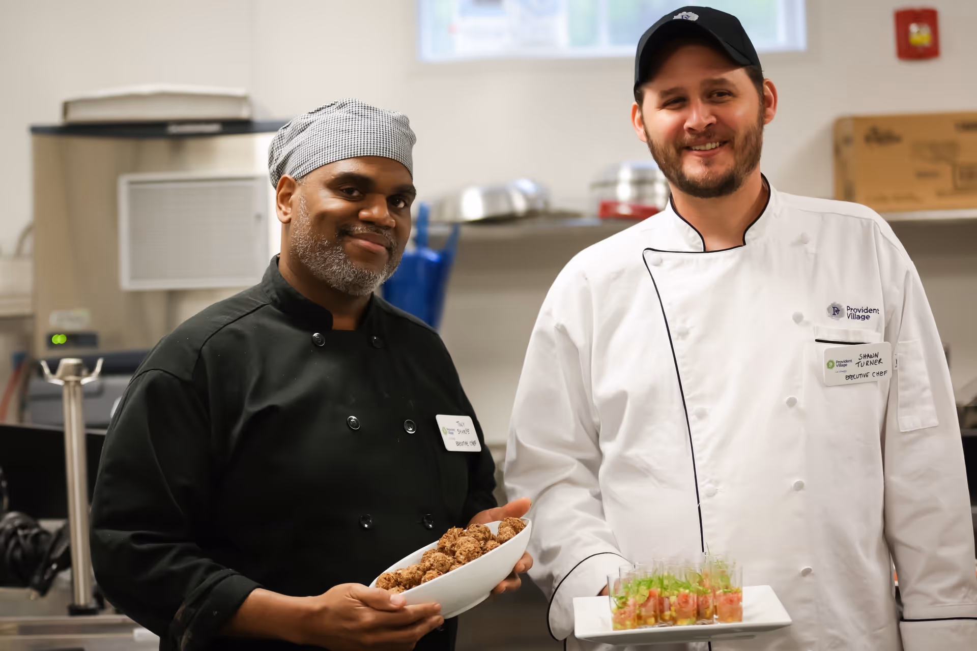 Two chefs standing in a kitchen, one wearing a black chef coat and checkered headscarf holding a bowl of meatballs, and the other wearing a white chef coat and black cap holding a plate with small appetizer cups.