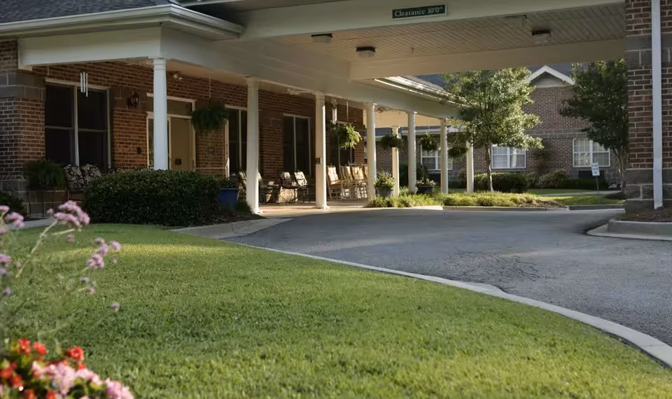 Covered driveway entrance to a brick senior living facility with white columns, rocking chairs on the porch, hanging plants, and well-maintained landscaping including green grass and flowering plants.