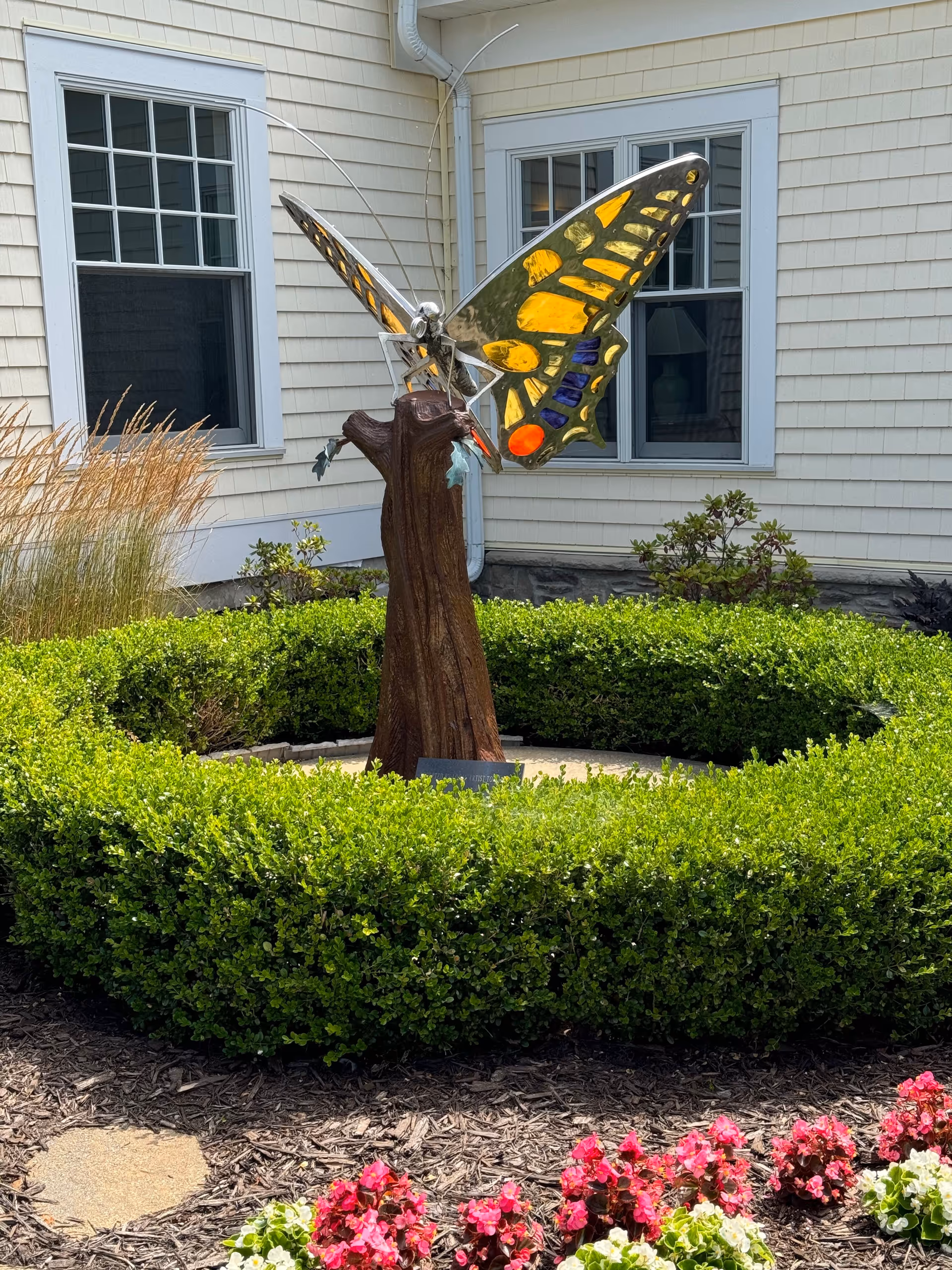 Butterfly sculpture on a wooden pedestal amid trimmed hedges in front of a beige house.