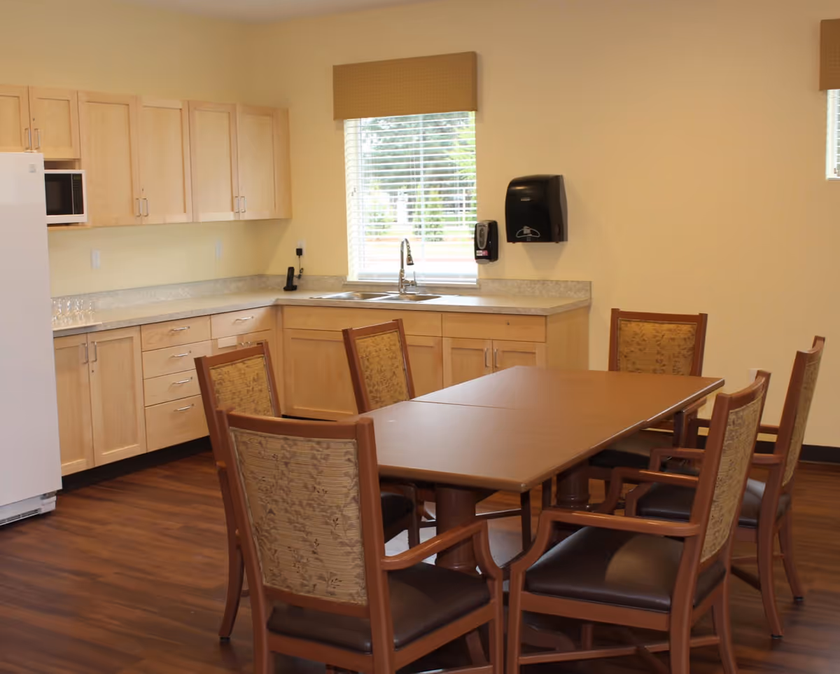 A dining area with a rectangular wooden table surrounded by six wooden chairs with cushioned seats and patterned backs. Behind the table is a kitchen area with light wood cabinets, a countertop, a double sink under a window with blinds, a white refrigerator, and a microwave. The floor is wooden and the walls are painted light yellow.