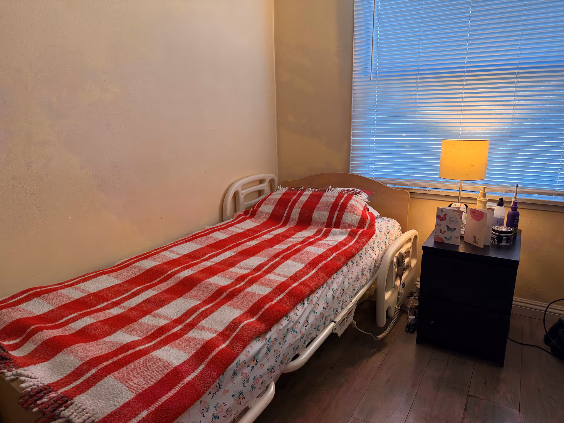 A hospital-style bed with a red striped blanket in small bedroom by a nightstand and lamp.