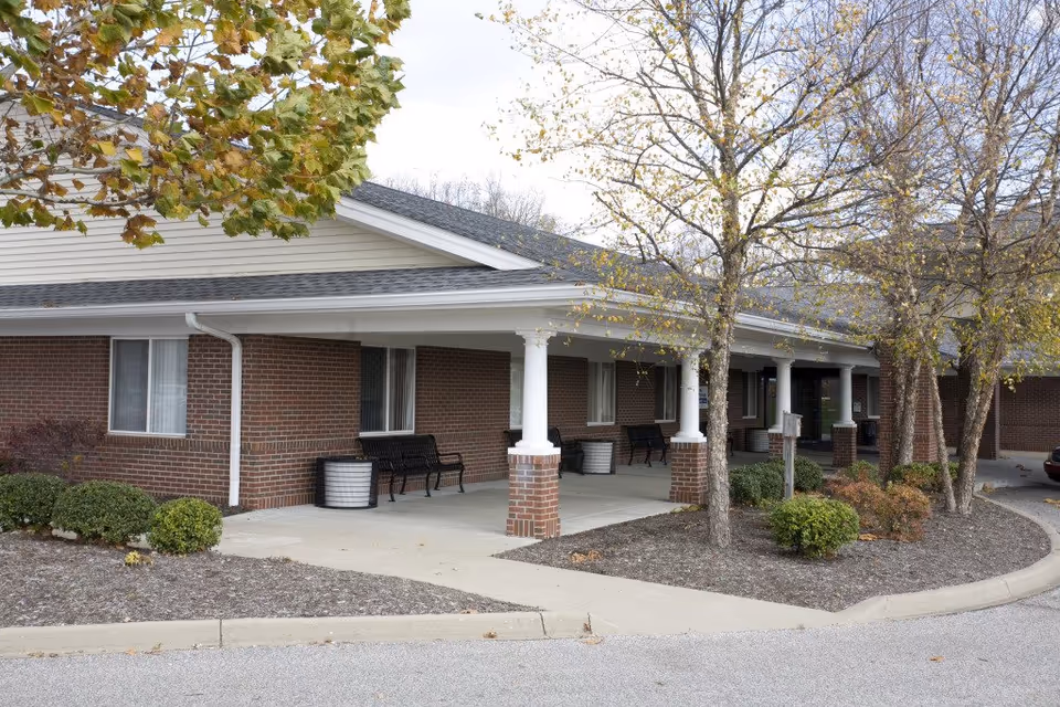 Exterior of a brick senior living facility with a covered porch, benches, and trees.