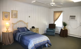 Spacious bedroom with a single bed, nightstand and lamp, recliner by a curtained window, and an air conditioner on white paneled walls.