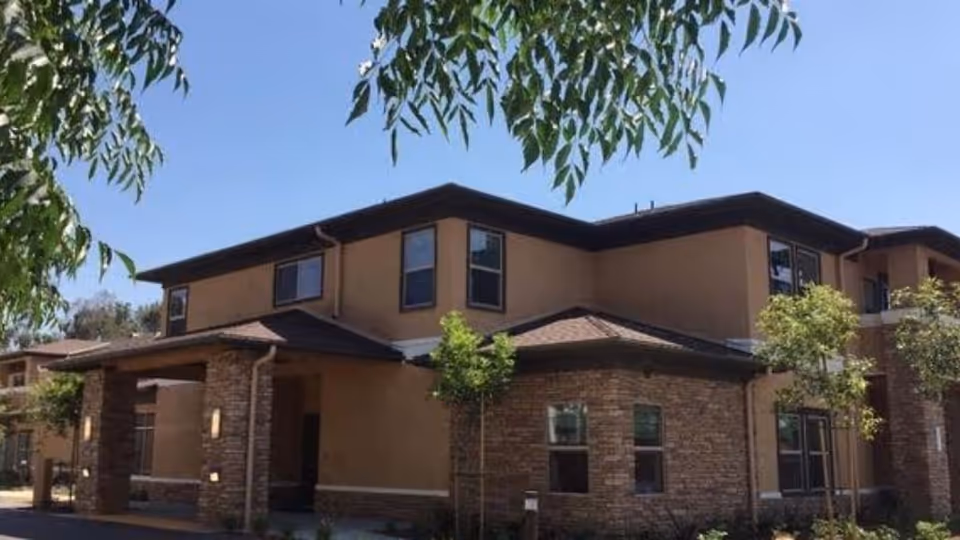 Two-story beige and brick senior living facility exterior with entrance canopy and surrounding trees.