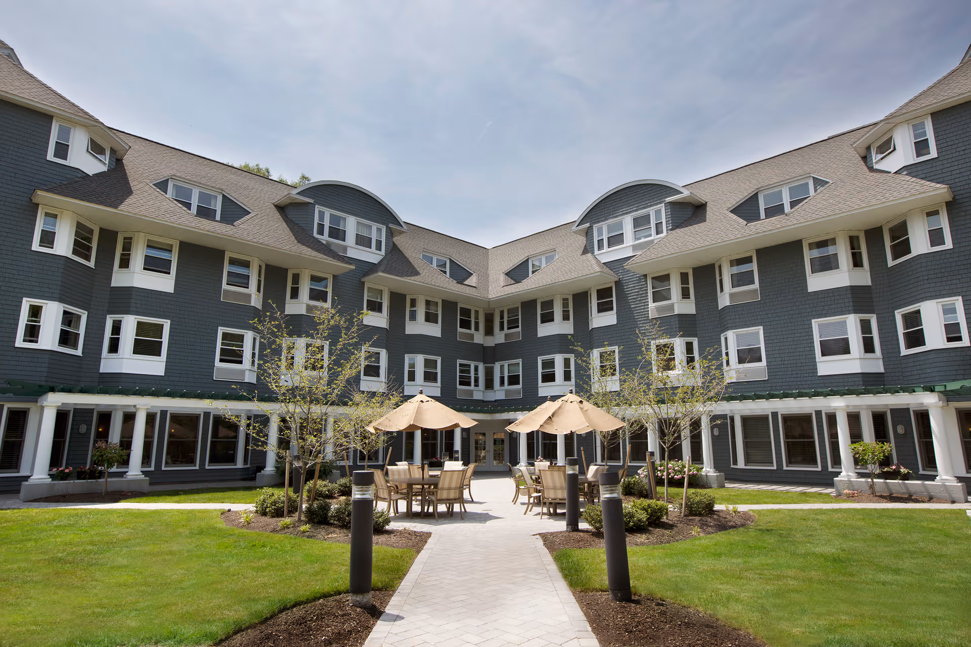 Courtyard with tables and umbrellas in front of a multi-story gray shingled senior living building.