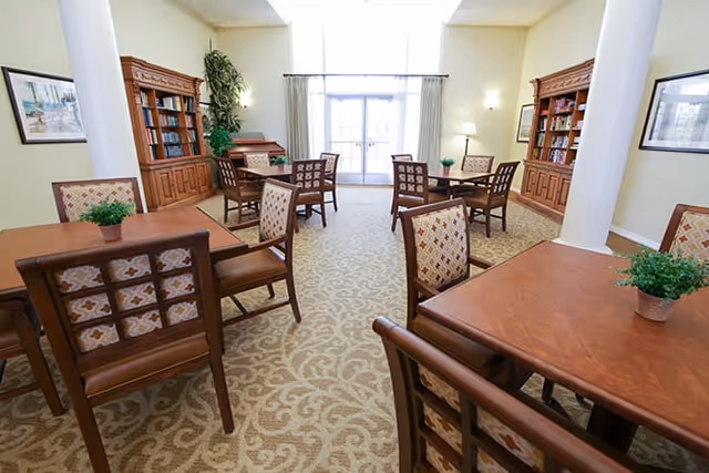 Bright communal room with wooden tables and upholstered chairs, bookshelves, plants, and large windows.