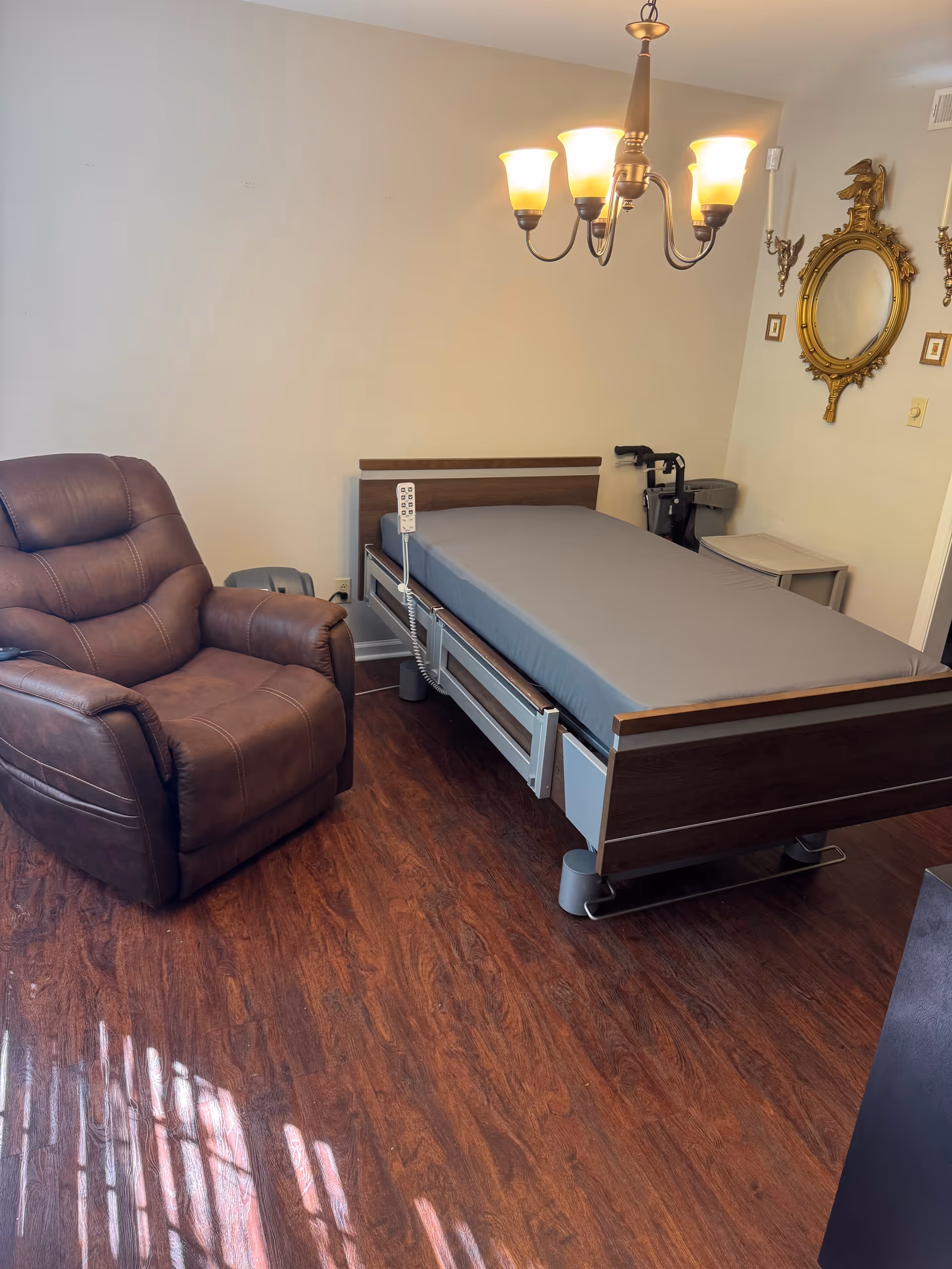 Senior living bedroom with adjustable bed, brown recliner, chandelier, mirror, and wood floors.