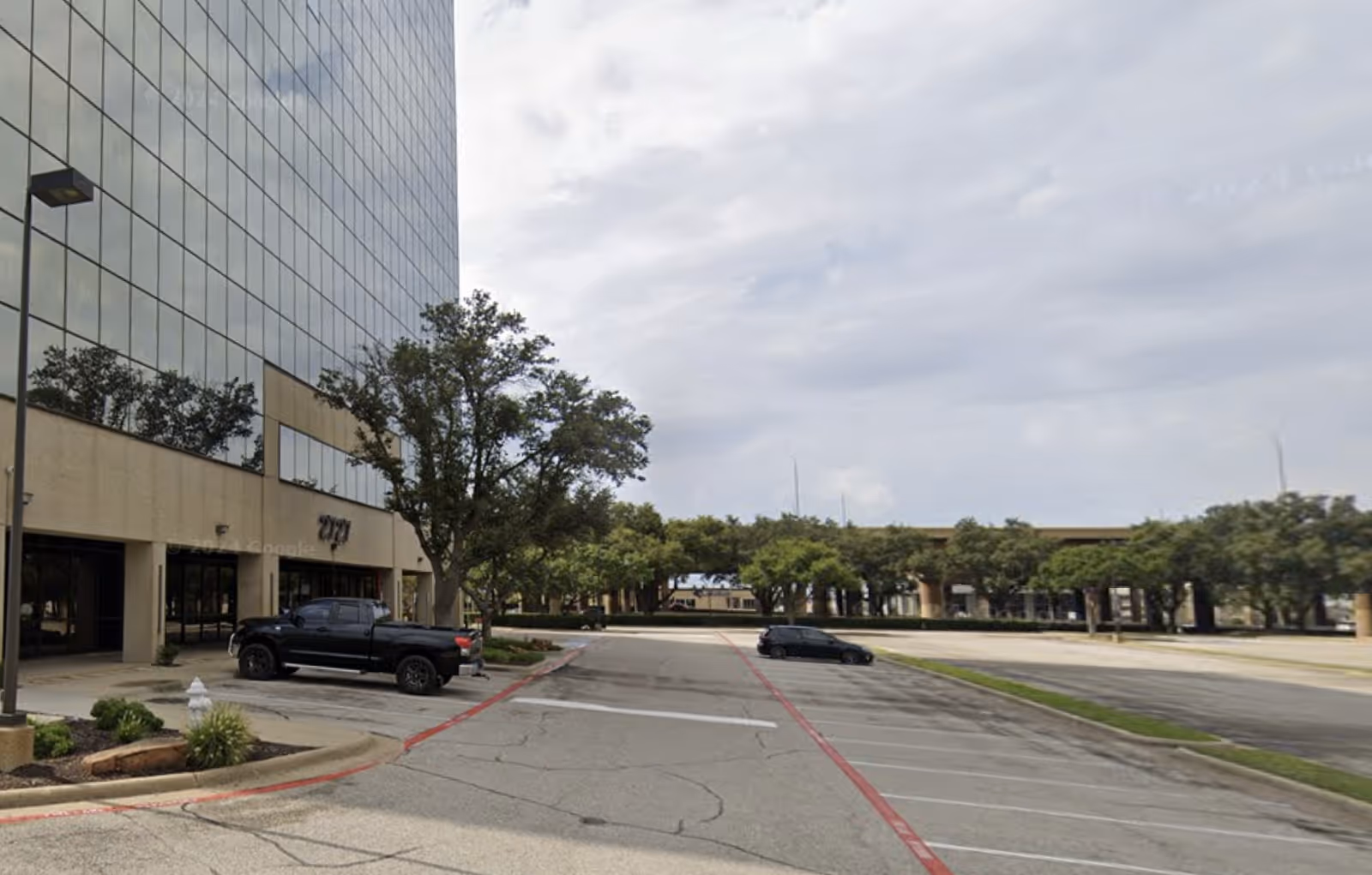 Outdoor parking lot beside a tall glass-building with trees and distant road.
