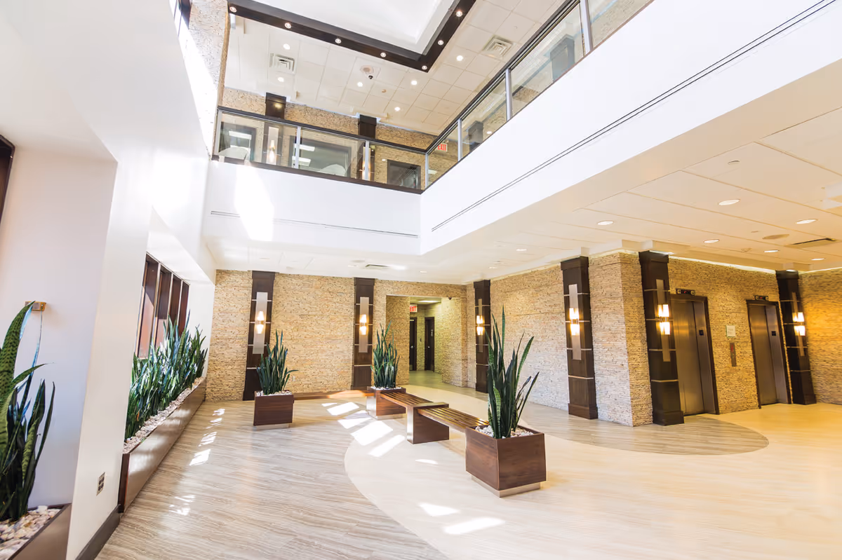 Bright, two-story modern lobby with benches, tall potted plants, and elevators.